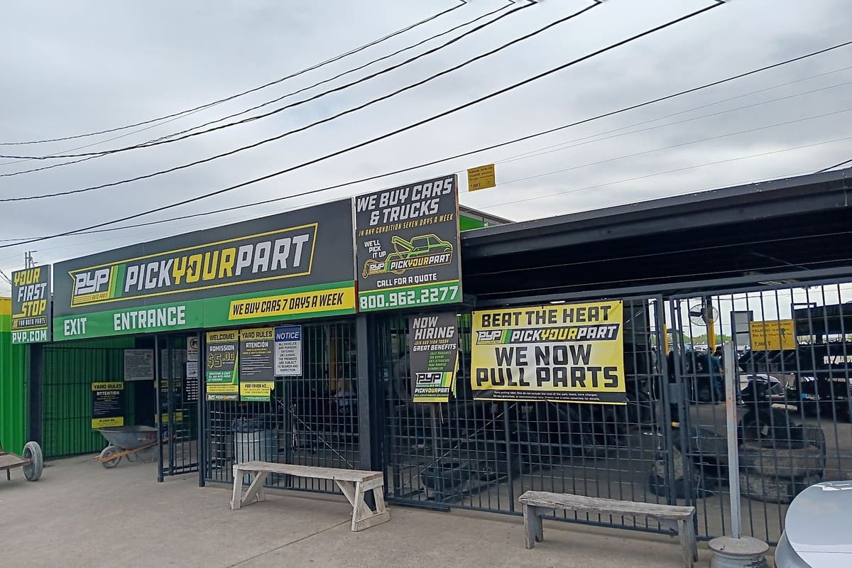 Pick Your Part - Houston Wallisville: Gated entrance and exit of a self-service auto salvage yard with green and black signs, benches and wheelbarrows out front