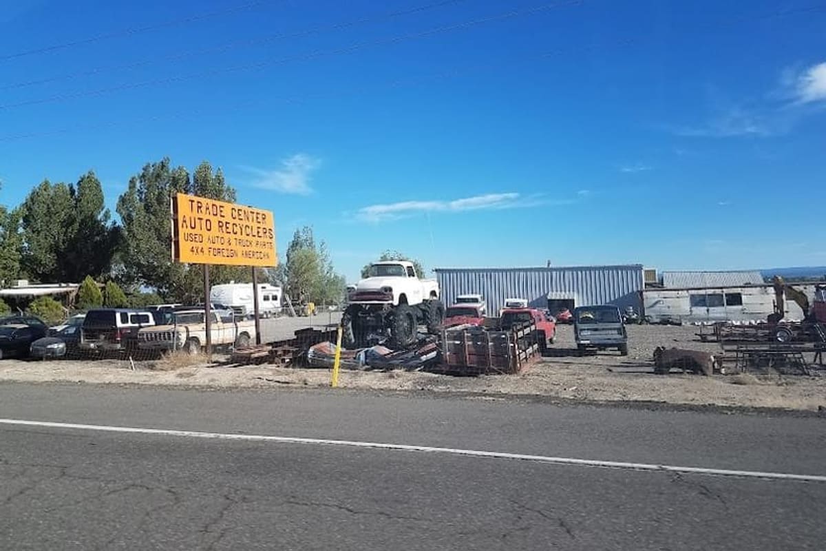 Trade Center Auto Recycling: Roadside salvage yard with an orange sign, a lifted white truck, stacked parts and vehicles, and metal buildings under a blue sky
