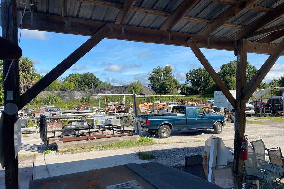 American Metal Salvage & Recycling: View from under a covered shed across a gravel salvage lot with a pickup truck, trailers, and scattered vehicles and parts