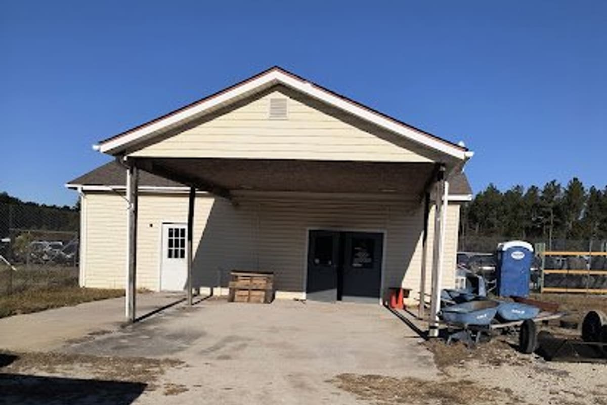 Foss U-Pull-It Jacksonville: Cream-colored single-story office building with a covered carport and double doors, set on a gravel lot under clear blue sky