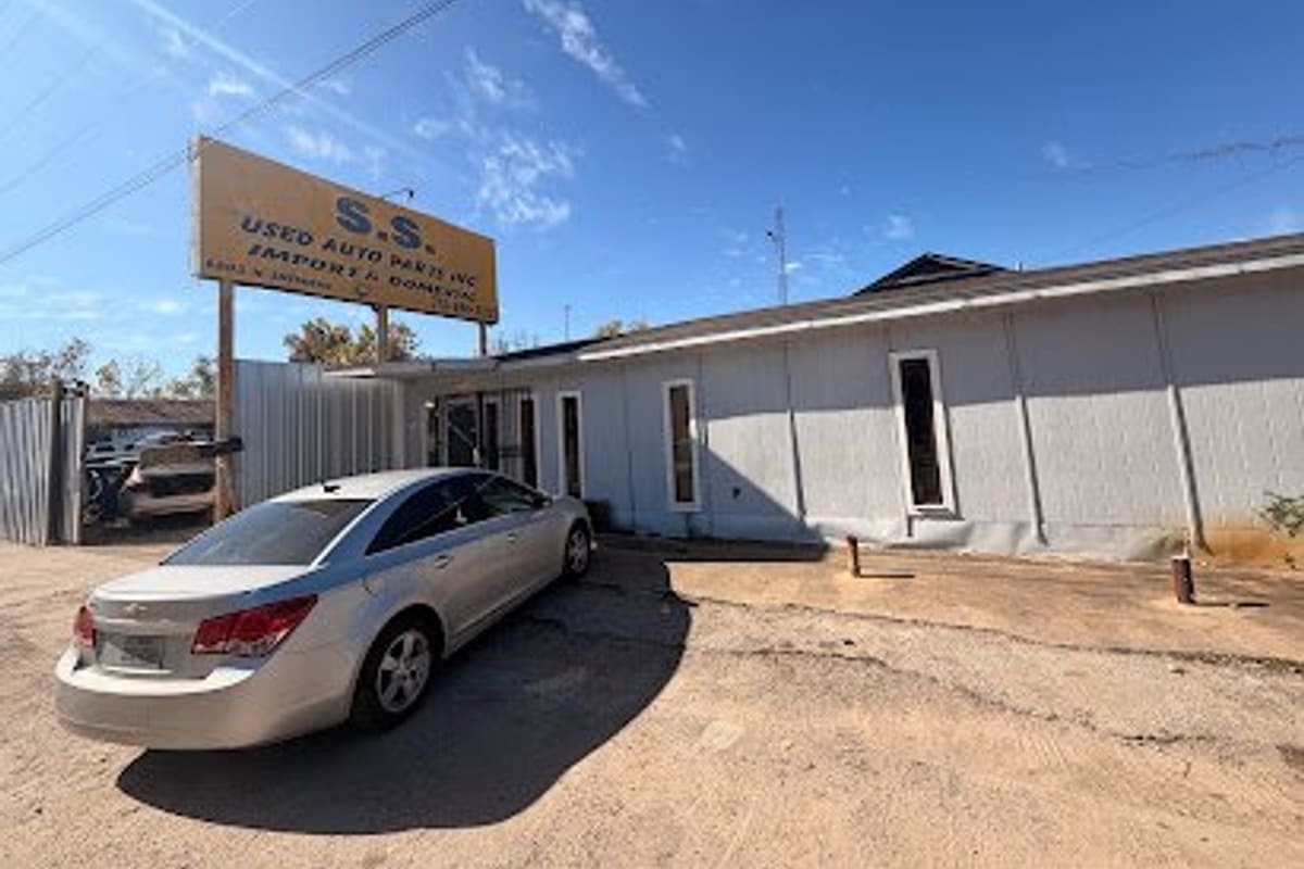 S S Used Auto Parts: Single-story gray office building with a tall yellow used auto parts sign beside a dirt lot and a parked silver sedan