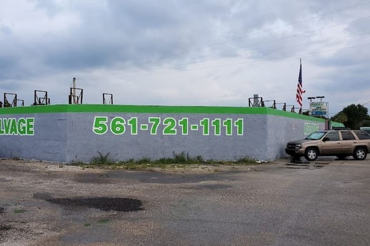 Ernies Affordable Auto Salvage: Gray perimeter wall painted with green lettering reading Auto Salvage and a phone number, with cars parked in the gravel lot