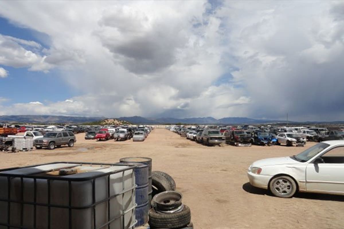 A 1 Auto and Truck Recyclers: Wide view of a dirt salvage yard with long rows of junked cars, tires and a tank in front, under a cloudy sky