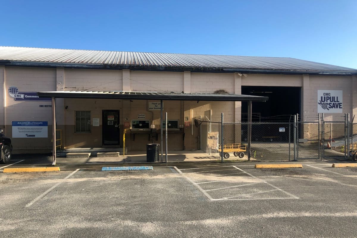 CMC Recycling: Single-story block building with a covered cashier window area, fenced yard gate and empty parking lot under a blue sky
