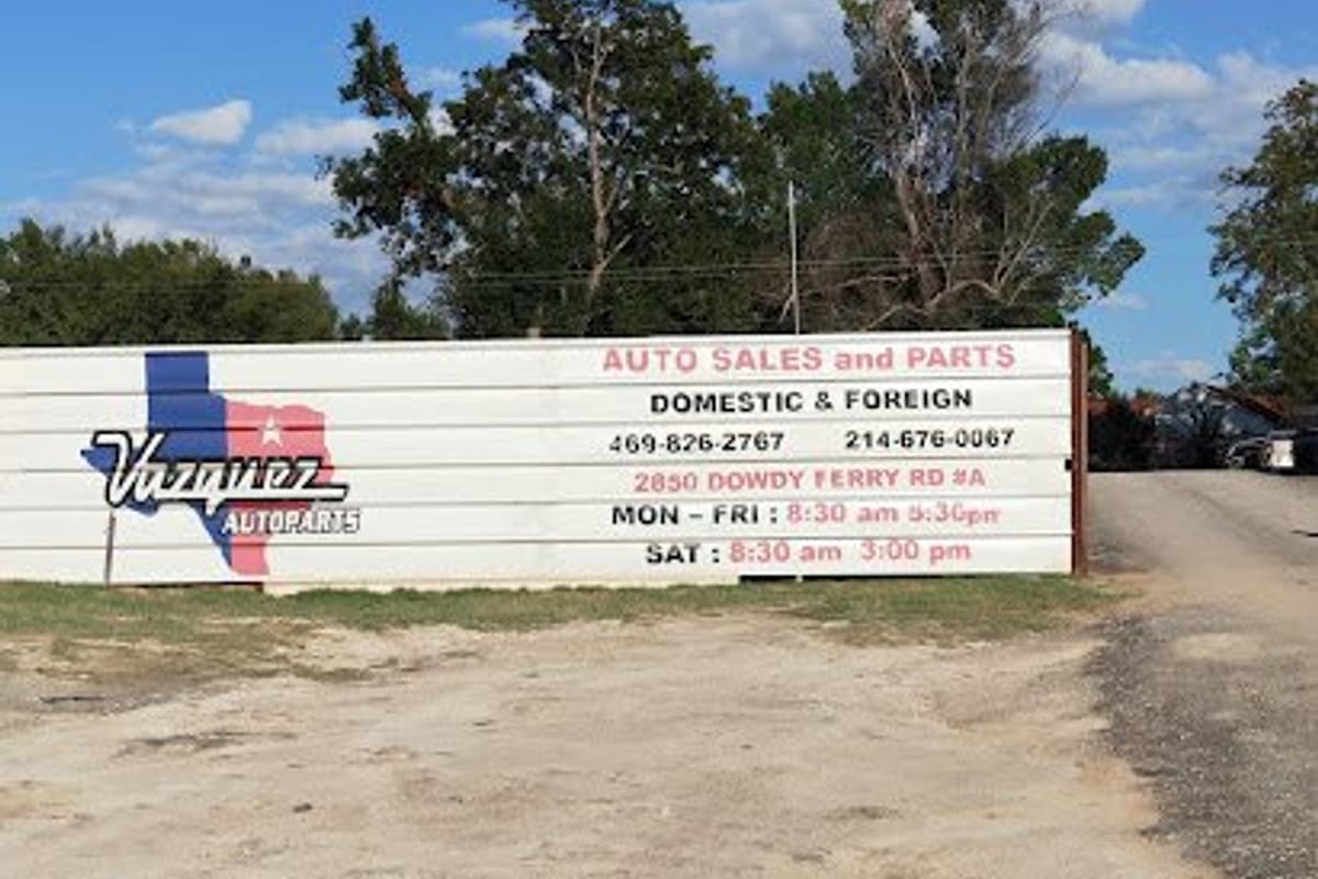 Vasquez Auto Parts and Sales: Large white roadside sign for auto sales and parts beside a dirt lot entrance under a blue sky with trees behind